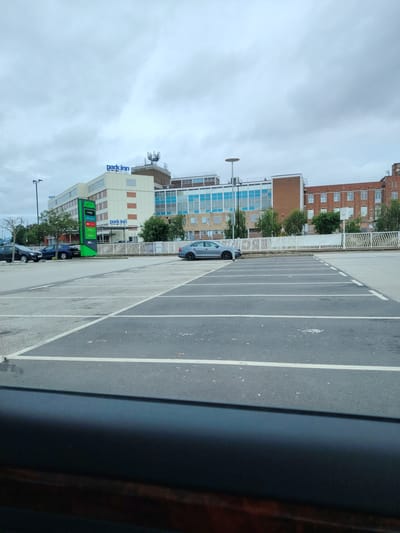 The image displays an outdoor scene under an overcast sky, captured from a vehicle, indicated by the dark, blurred obstruction in the lower foreground. A large, grey asphalt parking lot, sparsely occupied and delineated by white painted lines, extends from the foreground into the midground.
In the midground, a prominent green ASDA pylon sign stands centrally, listing services such as "George," "Click & Collect," "Groceries," "Kitchen," "Pharmacy," and "Opticians." To the left, several vehicles are parked, including a dark-colored SUV and a black sedan. A single grey sedan is parked further right within the marked spaces. A white, undulating fence runs horizontally, separating the parking area from the buildings in the background.
The background is dominated by multiple multi-story buildings. A building on the left prominently features "park inn" signage in blue lettering on its roof, characterized by light-colored panels and rectangular windows, some with orange-brown accents. Immediately behind and to the right of this structure is a larger building with a distinct blue-and-white facade, featuring numerous horizontal window sections. Further to the right, a building constructed of reddish-brown brick with multiple windows is visible. A rooftop structure with several telecommunication antennae is discernible on a building behind the "Park Inn" sign. Scattered green foliage and trees are visible between the fence and the buildings.
This scene is located in Peterborough, United Kingdom, specifically depicting the area of the Park Inn by Radisson Peterborough and the adjacent ASDA Peterborough Superstore at Rivergate. The photograph is taken from within the ASDA car park, facing the hotel and surrounding commercial structures.