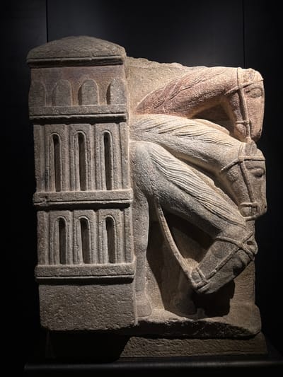 This is a stone relief sculpture depicting two horse heads, possibly part of a larger architectural element. The sculpture is carved in a rough, ancient style, with visible tool marks and wear. The horses' heads are rendered in profile, with their mouths slightly open and their manes indicated by parallel lines. They appear to be straining or in motion, with their bodies implied behind them. The lower portion of the sculpture features a decorative band with a series of horizontal grooves and raised sections, resembling a frieze or plinth. The material is a textured, light-colored stone, possibly granite or sandstone, with some reddish-brown discoloration on one of the horse's necks. The lighting creates strong shadows, highlighting the relief and texture of the carving. The sculpture is displayed against a dark, neutral background, suggesting it is an exhibit in a museum or historical site. Given the location context of Santiago de Compostela, Spain, this sculpture might be a fragment of Romanesque or medieval artwork, possibly from the cathedral or another significant building in the city. The style of carving and the depiction of horses are common in religious and secular art from the Iberian Peninsula during these periods. There is no visible text in the image.