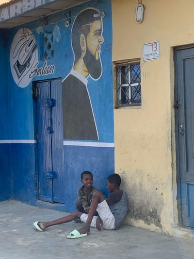 The scene depicts an outdoor street view in Hotoro South, Kano, Nigeria, featuring two male children in front of a building. The building's left section is painted bright blue with a white horizontal stripe, adorned with a large mural of a bearded man's profile and a circular "Saloon" logo featuring a barber's clipper. A blue double-leaf wooden door, secured with visible latches, is centered within the blue wall. The building's right section is a faded yellow-orange wall with significant staining and peeling paint near the base. This section includes a small, grilled window and a white rectangular sign above it, which reads "NO. 13 TSAMIYAR BOKA CITY OF KANO". A white outdoor light fixture is mounted above the sign. To the far right, a gray-blue door is partially visible.
Two male children are seated on the light gray concrete ground in the lower right foreground. The child on the left, younger, wears a camouflage long-sleeve shirt and dark trousers, and is crouched facing the other child, smiling towards the viewer. The child on the right, slightly older, wears a gray t-shirt with dark sleeves and white shorts, leaning against the yellow wall with his legs extended. He is wearing light green slide-on slippers. Both children are looking towards each other, engaged in interaction. The lighting indicates daylight conditions.