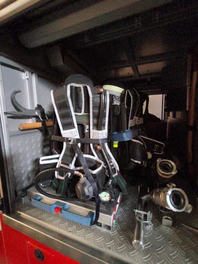 The image shows the interior of a fire truck compartment in Tukums, Latvia. The compartment is lined with a metallic diamond plate material. Dominating the center of the image are two self-contained breathing apparatus (SCBA) units, appearing to be of the MSA brand, with black harnesses and white reflective straps. One SCBA unit has a gauge visible. To the left of the SCBAs, mounted on the wall, is a metal hook with a wooden handle. Below the SCBAs, on a blue support, are two red and blue buttons. Towards the right of the frame, several metallic fire hose couplings are visible. The lighting is dim, suggesting the image was taken inside the truck.