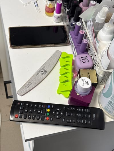 The image shows a cluttered white surface, likely a desk or shelf, with various personal care and electronic items. On the left, a black smartphone is placed horizontally. To its right, a light gray nail file with text indicating "GLOBAL FASHION®" and "MONOLIJSER PF-100/180" is positioned diagonally. A bright green, plastic toe separator lies to the right of the nail file. Further to the right, there is a collection of nail polish bottles, primarily in shades of purple and pink, some with black caps. One bottle has "KLIO" and "Color Base" visible. There is also a small white jar labeled "772 CUSHION GEL WHITE" sitting on a purple container. A gold-colored box with black text reading "m² nam" is visible behind the gel jar. In the background, a white plastic basket holds more items, including what appears to be a spray bottle and wooden sticks. Dominating the bottom of the image is a black television remote control with numerous buttons, including a power button, number pad, and various function keys. The remote has the model number "Y-72C3" printed on its lower right. A partially visible large white bottle with green and blue text is on the far right edge of the frame. The lighting suggests an indoor setting. There is no indication of the city of Емельяново, Russia, within the image itself.