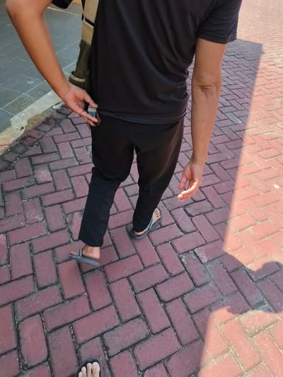 A person is walking on a brick-paved sidewalk in Barengkrajan, Indonesia. They are wearing a black t-shirt, black pants, and grey flip-flops. The person has their left hand in their pocket, holding something small. The sun is casting a shadow on the sidewalk.