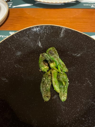Four blistered Padron peppers are arranged on a speckled dark plate. They have a shiny surface and are lightly charred in places. The plate sits on a wooden table next to a table setting with a fish-themed placemat. A white dish is partially visible.