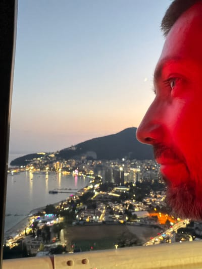 A close-up, profile shot shows the right side of a man's face, illuminated by a red light. He has dark hair, a beard, and is looking towards a city skyline at dusk. The city, identified as Košljun, Montenegro, is spread across a bay and hillside, with numerous lights twinkling from buildings and along the coast. The bay is dotted with boats, and the lights reflect on the water. A faint crescent moon is visible in the pale twilight sky. The foreground of the image features a blurry railing, suggesting the viewpoint is from a balcony, observation deck, or similar elevated structure. The overall impression is one of quiet contemplation against a vibrant urban backdrop.
