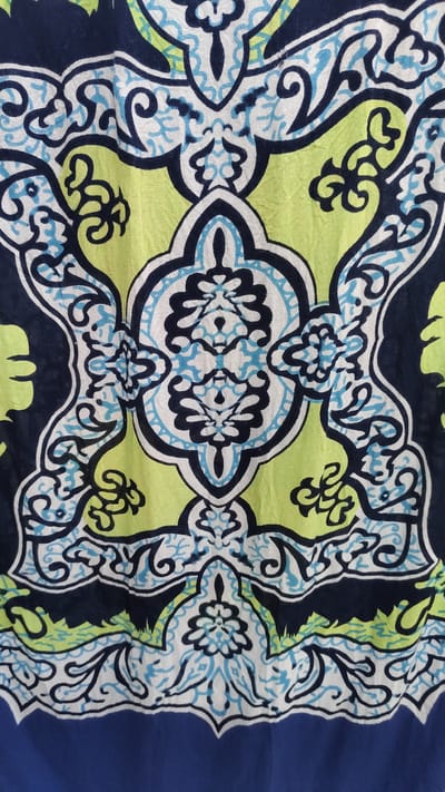 This is a close-up, full-frame shot of a fabric with a vibrant, intricate pattern. The pattern is predominantly in shades of navy blue, bright lime green, white, and light blue, with black outlines defining the shapes. It features symmetrical, ornate designs that resemble stylized floral motifs or mandalas. Swirling lines and petal-like shapes are characteristic of the design. The fabric appears to be slightly wrinkled, indicating it might be clothing, a tapestry, or upholstery. The overall impression is one of bold color and detailed artistry. No people, text, or specific setting elements are visible, other than the fabric itself. The location context of Gulistan, Uzbekistan, suggests this pattern might be representative of traditional Uzbek textile designs, which often feature complex geometric and floral motifs.