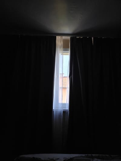 The image depicts a bedroom with dark curtains partially drawn, revealing a window with a view of buildings. The scene is dimly lit, suggesting either early morning or late evening. The focus is on the contrast between the dark interior and the light filtering through the window. The lower portion of the image shows a bed with white bedding. The buildings visible through the window appear to be residential with tiled roofs. The location is identified as Slivnitsa, Bulgaria.