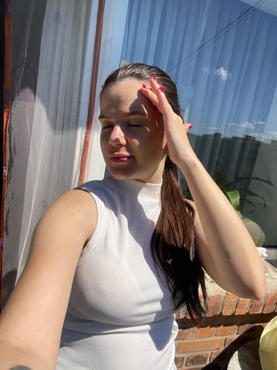A young woman with dark hair pulled back into a ponytail, wearing a white ribbed sleeveless turtleneck shirt, is standing outdoors, illuminated by bright sunlight. She has her right hand on her forehead, with her eyes closed and her head tilted slightly. A subtle sheen is visible on her forehead and nose due to the sunlight. Her fingernails are painted with bright pink polish. She is wearing a nose ring. Behind her, a sheer white curtain is partially visible, and beyond that, a large window reflects the blue sky with a single white cloud and the silhouettes of buildings. The scene appears to be on a balcony or rooftop, with a brick ledge in the foreground and a potted plant visible through the window. The overall setting suggests a warm, sunny day. The image was captured in Romny, Ukraine.