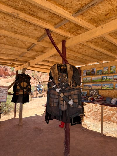 The image shows a tourist attraction in Tosor, Kyrgyzstan. Underneath a wooden shelter with exposed beams and particleboard roofing, two sets of traditional Mongolian armor are displayed on posts. The armor is dark leather with fur trim and metal accents. Above the armor, small landscape paintings and souvenirs are arranged along the top of the shelter. In the background, a man is sitting on a raised wooden platform. A sign with information about archery and photo opportunities is visible in the lower right corner. The ground is reddish-brown soil.