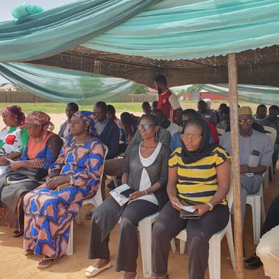 This media file captures an outdoor gathering in Unguwar Saya, Nigeria, likely during the daytime. The scene appears to be a community event or meeting, with a group of people seated on white plastic chairs under a makeshift canopy. The canopy is constructed with a corrugated metal roof covered by brown fabric, supported by wooden beams, and draped with light blue and green fabric curtains.
The subjects are diverse, consisting of men and women of various ages. Many individuals are dressed in traditional Nigerian attire, characterized by vibrant patterns and colors, such as the striking blue and orange geometric print worn by a woman in the foreground. Others are in more casual Western-style clothing. Some people are looking forward, suggesting they are attending to a speaker or presentation, while others have their eyes closed or are looking down, possibly due to the heat or fatigue.
Notable details include the presence of cell phones, with one woman holding a smartphone and another holding documents. A flyer is visible in the foreground with some text, but it is not clearly legible. The ground is covered in reddish-brown earth, and in the background, brick structures and some greenery can be seen, hinting at a rural or semi-urban setting. The overall mood seems to be one of community engagement, with a sense of focus among the attendees. The time of day appears to be late morning or afternoon, given the sunlight and shadows. The weather seems to be warm, as indicated by the attire and the need for shade.