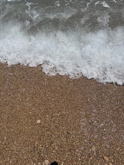 The image shows a close-up view of the shoreline at a beach. On the left side of the image, white foamy waves crash onto the shore. The right side consists of a tan-colored pebbled beach, with varying sizes of small rocks and pebbles. The pebbles near the water appear wet and reflective. The location is Vinaròs, Spain. No people are visible in the scene.