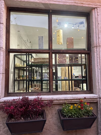 The image shows a window display in Domingo Arenas, Mexico. The window is divided into panes, with the left panes displaying stone slabs and light fixtures. The right panes show a shelf system displaying various stone pieces, including a cartoon cat figurine, and price tags indicating "$30 MXN." Plants are displayed in planters outside the window on the right, with red and green foliage. The wall surrounding the window is textured. The OCR in the window displays the words "Proudly Made in Mexico" and "Orgullosamente Hecho."