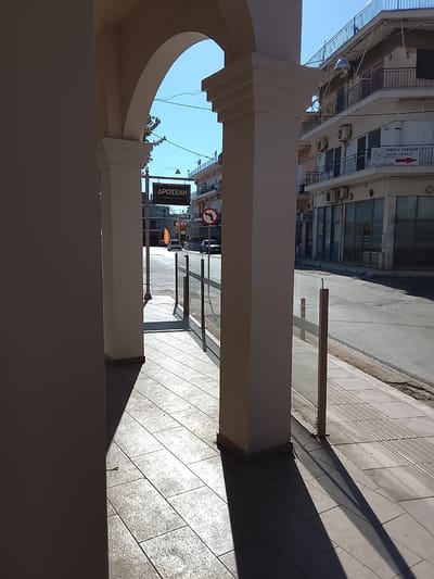 A sunlit, angled view from beneath a colonnade in Λεχαινά, Greece reveals a street scene. The colonnade's columns cast stark shadows across the tiled pavement. Beyond the colonnade, a street displays signage, including "ΔΡΟΣΕΛΗ" and a "no left turn" traffic sign. Cars are parked along the road. Buildings line the street, one displaying the sign "VASSILIS PANTAZIS AGROFRUIT." The scene is bathed in bright daylight under a clear, blue sky. There is no visible human activity.