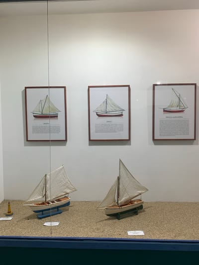 The image shows a museum display with three framed pictures of sailboats hanging on a white wall. Below the pictures are two model sailboats placed on a sandy surface, with a small lighthouse to the left of the first sailboat. The framed pictures are labeled "OREJETA", "ZANCO", and "PIRAGUA MARGARITEÑA". The display is behind a glass panel, as indicated by reflections. The overall environment suggests an indoor exhibition space dedicated to maritime history or local vessels in Boca del Río, Venezuela.