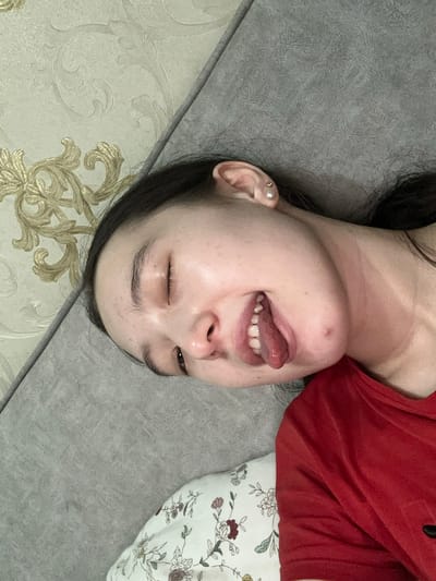 An individual with closed eyes, dark hair, and fair skin lies on a textured gray surface, possibly a sofa, in Nookat, Kyrgyzstan. The individual wears a red t-shirt. Their mouth is open, with their tongue extended. A patterned pillow is visible beneath their head. The background features wallpaper with a floral design.