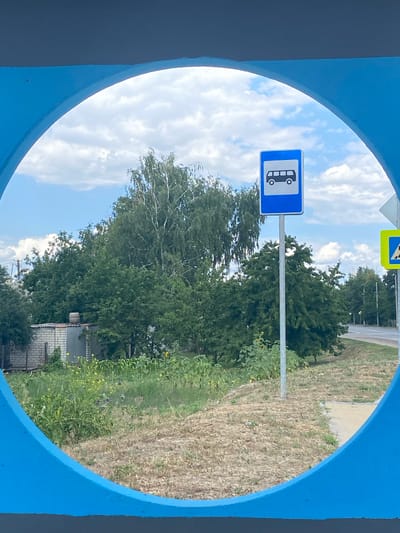 The image shows a bus stop sign on a pole against a backdrop of trees and a cloudy sky. The sign is blue and white with a black silhouette of a bus. To the right of the bus stop sign, a yellow and black pedestrian crossing sign is visible. The foreground is dominated by a bright blue, circular frame. In the background, there are trees, a small brick building, and a road. The scene appears to be a roadside location in Shulhynka, Ukraine, likely a rural or suburban area given the presence of vegetation and a modest building. There are no people visible in the image.
