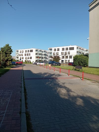 The image depicts a modern apartment complex in Radzymin, Poland, under a clear blue sky. The buildings are white with dark-trimmed windows and balconies. In the foreground, a paved walkway with red bollards leads towards the buildings, with parked cars visible in the distance. A park bench sits on a grassy area next to the walkway. The scene suggests a residential area with ongoing development or upkeep, evidenced by the construction or landscaping elements.