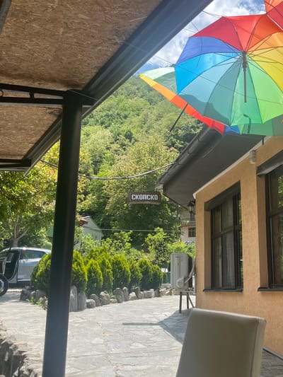 This image depicts an outdoor setting in Durachka Reka, North Macedonia, viewed from beneath a covered patio. The foreground displays the underside of a light-colored OSB panel roof, supported by dark metal structural beams and a cylindrical post on the left. To the right, a building with a textured light brown stucco-like facade features multiple dark-framed windows and a grey rain gutter along its roofline. Several open, rainbow-colored umbrellas are suspended decoratively beneath the building's eaves. An outdoor air conditioning unit is positioned on a metal stand beside the building.
The mid-ground features a stone-paved patio area. A line of small, conical evergreen shrubs, bordered by natural rocks, separates the patio from a parked silver SUV with its driver's side door open, located on the left. In the background, a sign with a brown frame and white Cyrillic text, reading "СКОПСКО" (SKOPSKO), hangs amidst lush green deciduous trees. Densely forested mountains ascend in the distance under a partly cloudy sky. The backrest of a light-colored chair is visible in the lower right corner. The scene is illuminated by bright daylight, casting clear shadows. No individuals are present.