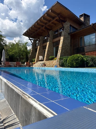 The image depicts a modern, multi-story building with a rustic architectural style, characterized by exposed wooden beams and stone accents, situated in Dobrinishte, Bulgaria. In the foreground, a bright blue swimming pool with tiled edges reflects the clear sky and the building. Several wooden lounge chairs and a white umbrella are arranged near the pool, suggesting a leisure area. Lush green trees and manicured bushes surround the property, contributing to a natural and serene environment. A blue pool skimmer lies on the ground near the building, indicating recent or ongoing maintenance. The overall scene captures a luxurious outdoor setting, likely a private residence or a resort, under a bright, partly cloudy sky.