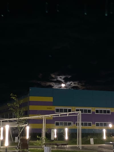 The image captures a night scene in the city of Pokhvistnevo, Russia. A modern, multi-story building with brightly colored horizontal bands of purple, yellow, and teal dominates the mid-ground. The building features numerous rectangular windows. In the foreground, a white, open-air structure with vertical supports and a horizontal roofline is visible, along with several tall, glowing vertical lights that resemble bone shapes. There are also glimpses of trees and outdoor seating elements. The upper portion of the image is filled with a dark, cloudy night sky, through which the moon is partially visible, creating a luminous glow behind the clouds.