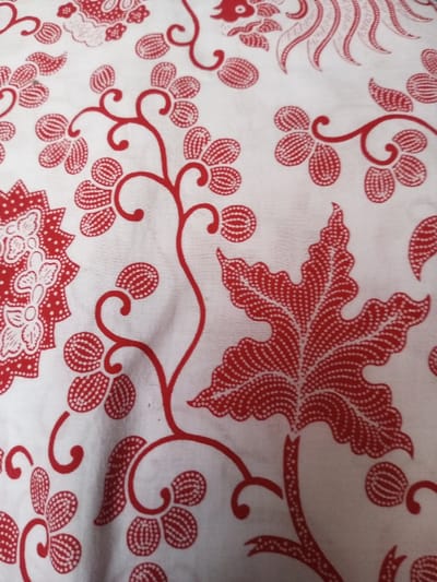 The image showcases a white fabric adorned with a red batik pattern. The design includes stylized flowers with clustered petals, winding stems, and a prominent leaf with detailed veins. Each red element consists of numerous small dots, creating a textured appearance against the white background. The overall pattern suggests a traditional or ethnic textile design.