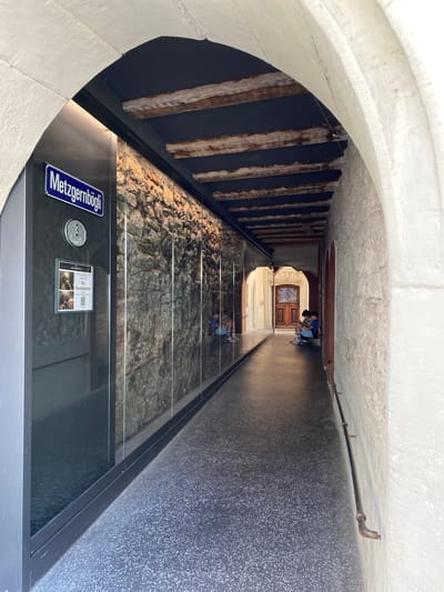 This image depicts a narrow, arched passage or underpass, likely in an urban setting. The perspective looks along the length of the passage, which features a modern glass floor covering an excavated archaeological site. Beneath the glass, rough stone and rock foundations are visible, suggesting ancient structures.

On the left side of the passage, a wall incorporates exposed wooden beams. The right wall is dark and textured, with a metal pipe running along its upper edge. The arched ceiling is made of a light-colored, rough-textured material.

Towards the far end of the passage, two individuals are present. One person, possibly a child, is bent over, seemingly observing something on the ground or within the glass display. Another person stands nearby. Beyond them, an ornate wooden door and an arched doorway are visible, leading into another area.

In the foreground, a blue street sign reads "Metzgernbögli." Adjacent to it is a circular silver sign with the number "3" and the partially visible text "Weimarkt." An information panel is also present, featuring images, a QR code, and text including "Der Gewölbekeller" (The Vaulted Cellar).

The scene suggests a historical exhibition or display integrated into a public thoroughfare, allowing visitors to view archaeological remains while navigating the passage. The lighting indicates daytime, likely within a sheltered or indoor environment. The presence of street signs and information panels indicates a publicly accessible site. The visible text and architectural style point to a location in Lucerne, Switzerland.