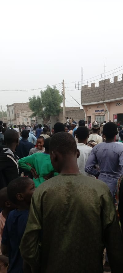 This image captures a dense gathering of people outdoors, likely in a street or open area, under an overcast or hazy sky. The perspective is from within the crowd, looking forward, with many individuals visible from behind.

In the immediate foreground, the back of a young person wearing a dark olive-green long-sleeved garment with a subtle circular pattern is prominent, occupying much of the lower-right portion of the frame. To their left, another young person in a dark blue shirt is partially visible from the back.

Further into the mid-ground, amidst the crowd, a young boy in a bright green long-sleeved shirt stands out, facing slightly towards the left and looking towards the camera with a clear smile. His hands are on his hips. Other people of various ages, predominantly male, are visible, many wearing light-colored traditional garments, while some are in darker clothing. A person in a light blue striped top is seen towards the right mid-ground.

The background features several buildings, suggesting an urban or semi-urban environment. Some buildings appear to be residential, while one on the far right has a commercial front. This building is constructed with light-colored concrete blocks and has a distinctive crenellated top edge. A sign on this building reads "BASHIR" in large blue letters, with smaller, lighter text underneath that appears to say "Super and Eggs" or "Super n Eggs". Another building further to the left is older, with a lighter facade.

An electrical pole with numerous wires runs diagonally across the mid-ground, and a large green-leafed tree is situated between the pole and the buildings on the left. More people are gathered around the base of the tree and near the buildings. The overall impression is one of a busy community event or a bustling street scene. The hazy sky indicates either an overcast day or atmospheric conditions reducing visibility.