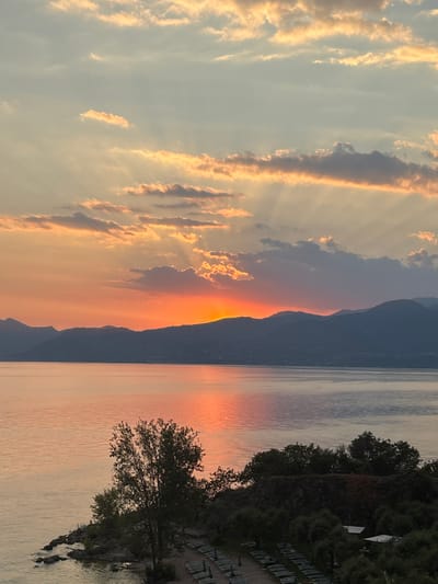 The image captures a picturesque sunset over Lake Garda, Italy, from a viewpoint on the western shore. The sun, partially obscured by clouds, casts a warm, orange glow across the sky, illuminating the scattered clouds with hues of yellow and pink. Rays of light beam through the clouds, creating a dramatic effect. Below, a range of dark, silhouetted mountains forms a striking backdrop against the vibrant sky. The calm waters of the lake reflect the colorful sunset, creating a shimmering, warm-toned surface. In the foreground, a small, rocky peninsula juts into the lake, covered with lush green trees and vegetation. Several lounge chairs are visible on a sandy area, suggesting a recreational space. The overall scene evokes a serene and tranquil atmosphere.