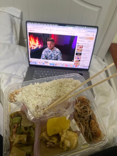 The scene is an interior view, likely a bedroom in Huangdao District, China, displaying a meal in conjunction with a laptop. A silver laptop rests on white bedding, open to a YouTube video. The video features a medium shot of a male subject with a beard, wearing a patterned shirt, against a background depicting a digital fireplace on the left and a wall with red and purple lighting on the right. In the foreground, a clear plastic multi-compartment takeout container is presented. The largest compartment contains white rice, partially mixed with brown rice. A lower-left compartment holds green vegetables with a sauce. The central lower compartment contains pieces of chicken with diced yellow vegetables in a yellowish-brown sauce. The lower-right compartment holds a stir-fried dish with noodles, bean sprouts, and other ingredients. A pair of wooden chopsticks rests across the rice compartment. White bedding surrounds the laptop, and a white headboard or wall paneling is visible in the background, with a dark brown surface on the far right.