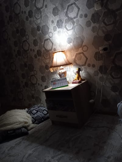 This image depicts a dimly lit bedroom interior, possibly in an apartment or house located in Әуезов ауылдық округі, Kazakhstan. The focal point is a bedside table with a lamp casting a warm glow onto the hexagonal-patterned wallpaper. On the table sits a small calendar, a decorative vase, and a yellow stuffed animal wearing a hat, positioned on a red object. A power outlet and a light switch are visible on the wall. To the left of the table, two pillows are placed on what appears to be a bed or couch, one with a textured white cover and another with a black and white geometric pattern. The floor is covered with a textured, gray carpet. The overall scene suggests a domestic setting, likely in the evening or night, given the artificial lighting.