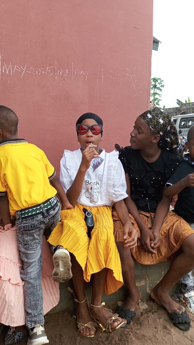 The image depicts a casual outdoor scene featuring four young individuals, likely children or teenagers, seated against a plain reddish-brown wall in Agburu Uke, Nigeria. The setting appears to be daytime, with bright, even lighting.
The central figure is a girl with dark skin, wearing a white ruffled blouse, a long yellow pleated skirt, and gold ankle-strap sandals. She is adorned with red sunglasses and a black crocheted cap, and a long, beaded necklace hangs around her neck, featuring a small pouch or bag. She is actively drinking from a clear plastic sachet, possibly containing a beverage, using a straw.
To her immediate right, another girl, also with dark skin, wears a black top with embellishments, a black and gold patterned headscarf, a brown or tan pleated skirt, and black floral slide sandals. She is looking towards the central girl with a calm, slightly smiling expression. On the far left, partially obscured, is a child in a bright yellow polo shirt and faded blue jeans, notable for a distinctive green, white, and black patterned belt that reads "TRAIN TEAM." Only the head and shoulder of a fourth child in a black top are visible on the far right.
The ground beneath the individuals is dusty or sandy. In the background, beyond the wall, a white vehicle and some sparse vegetation can be vaguely seen.
Notable text is written on the wall in what appears to be chalk or light paint, stating "MAY-2025" and "NGA DAYS 1-24-36." On the central girl's white blouse, "Bech" and a graphic are visible.