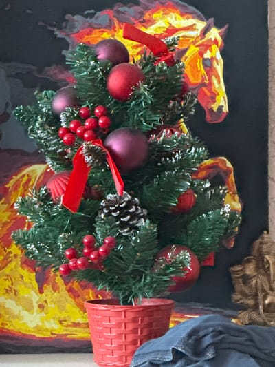 The image shows a miniature Christmas tree adorned with red ornaments, berries, pinecones, and red ribbons. The tree is placed in a red woven basket. In the background, a painting depicts a horse with a fiery mane and tail against a dark background. To the right of the tree, a golden statue, possibly of a deity, is visible. A dark blue cloth is draped in the foreground. The scene is indoors, likely in a residential setting. There is no discernible information in the image that would indicate its location in the city of Tambov, Russia.
