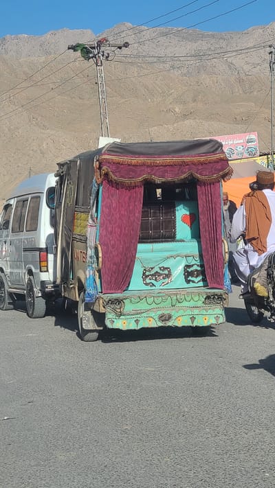 The image shows a decorated rickshaw with a maroon and gold fringe canopy. The rickshaw body is painted turquoise with decorative designs. A man wearing a brown and orange robe with white pants and a brown hat is visible. The rickshaw is parked on a paved road in Bostan, Pakistan. Power lines and a mountain are visible in the background.