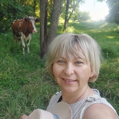 A woman with blonde hair and blue eyes is taking a selfie in a grassy, tree-lined area. She is wearing a striped top and holding a straw hat. Behind her, a brown and white cow is standing in the grass. The scene appears to be outdoors, possibly in a rural or park-like setting, with sunlight filtering through the trees. The location is identified as Voronkiv, Ukraine.