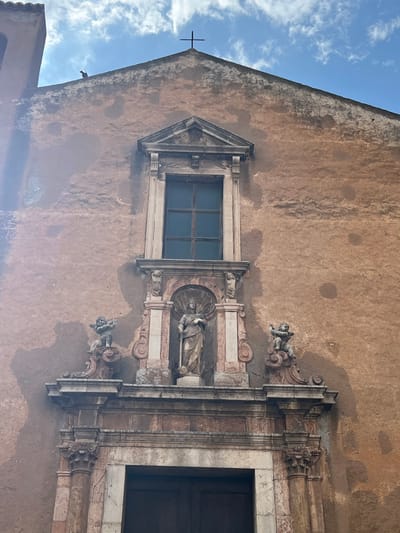 The image shows the facade of a church in Taormina, Italy. The building is made of a reddish-brown stucco material and has a large, dark-colored door at the bottom. Above the door is a decorative architectural element featuring columns and a central niche. Within the niche stands a statue of a robed figure holding a staff. Flanking this central element are two smaller sculptures of cherubic figures. Above the niche is a rectangular window with a dark frame. The entire facade is crowned by a pediment with a cross at its apex. The sky above is blue with scattered white clouds. A bird is perched on the roofline of the church.