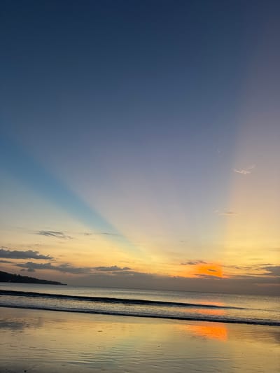 This is a photograph of a sunset over the ocean. The sky is a gradient of deep blue to bright orange and yellow, with some wispy clouds. Sunbeams are visible, fanning out from the horizon. The ocean is calm, with gentle waves lapping at the shore. The wet sand reflects the colors of the sky. The scene evokes a sense of peace and tranquility. The time of day is sunset, and the weather appears to be clear. The location is Kuta Selatan, Indonesia. There is no visible text in the image.