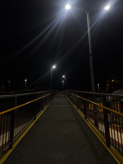 The image shows an elevated pedestrian walkway at night in Bassendean, Australia. The walkway is flanked by dark railings on both sides, painted yellow on the inner part. Streetlights illuminate the walkway. In the distance the path appears dark and the view is straight along the path.