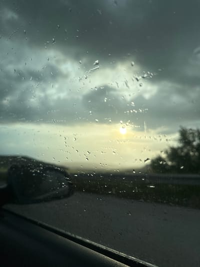 The image appears to be taken from inside a vehicle, possibly a car, looking through a rain-covered window. Outside, the weather seems overcast, with dark, stormy clouds dominating the sky. A blurred view of trees and a side-view mirror of the vehicle are visible. A bright spot in the distance suggests a break in the clouds or the sun attempting to shine through. The image does not depict Kipra, Bulgaria, nor any other identifying location. No people are visible in the image.
