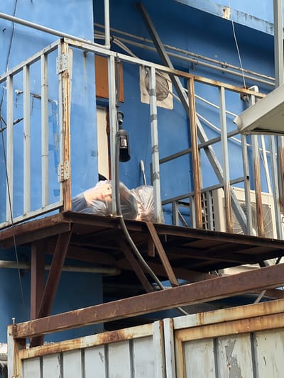 This image depicts an exterior scene in Gangseo-gu, South Korea, characterized by a weathered blue building and multiple rusted metal structures.
A prominent feature is an elevated, heavily rusted metal platform supported by equally rusted metal beams. On this platform, two clear plastic bags are visible. The left bag contains the head and shoulders of what appears to be a mannequin or doll, featuring light skin and dark hair/wig. A second similar bag is positioned to its right. A black cylindrical object, possibly a lamp or lantern, hangs from a vertical metal bar that is part of a blue-painted railing system enclosing the platform. This railing exhibits significant rust and peeling blue paint. A white-handled item is partially obscured behind the right plastic bag.
The building wall behind the platform is painted a vibrant blue, showing signs of wear and peeling paint. Multiple silver-colored pipes run horizontally across this blue wall. To the right, two outdoor air conditioning units are mounted against the blue wall, below the level of the platform. A rectangular brown panel is affixed to the blue wall above a white doorframe or window frame, behind the left side of the platform. A circular vent or similar structure is partially visible higher on the blue wall, to the right of the brown panel.
Below the main platform and its supports, a section of a heavily rusted, off-white or light grey metal gate is visible, displaying extensive paint chipping and rust. The overall scene suggests an urban or industrial environment with visible signs of aging and exposure to the elements. No individuals are present, and no actions or interactions are occurring.