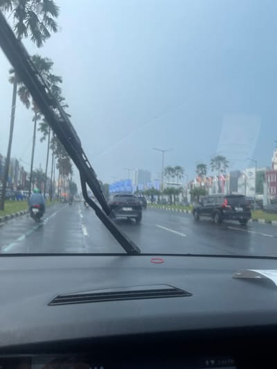 The image depicts a view from inside a vehicle looking through a wet windshield on a cloudy day. The windshield wipers are partially visible in the foreground. The scene outside shows a wet road with several vehicles, including cars and a motorcycle with a rider wearing a blue jacket and green helmet. Palm trees line the left side of the road, interspersed with buildings. The road appears to be a multi-lane street with traffic moving in the same direction. The environment suggests a tropical or subtropical urban setting, likely during or after rainfall. The presence of palm trees and the architecture of the buildings are consistent with locations in Indonesia, such as Pakulonan Barat. The time indicated at the bottom right appears to be 2:54 PM. A red rubber band is visible on the dashboard of the car.