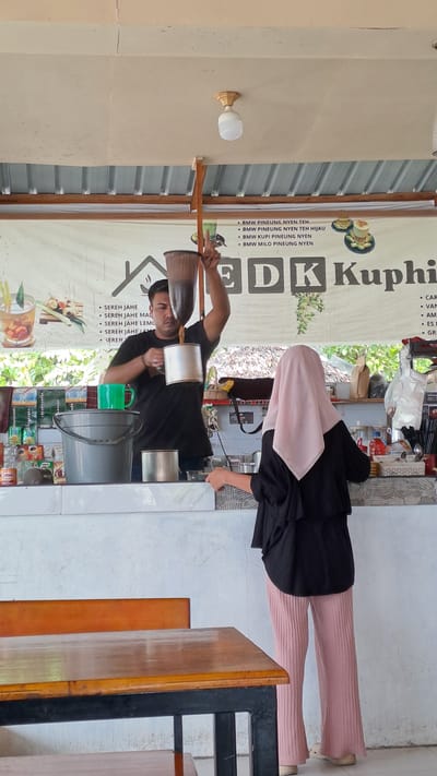 This media file, taken in Julikeude Dua, Indonesia, captures a scene at an outdoor food or drink stall. Two individuals are visible: a man in a black t-shirt appears to be preparing a drink, holding two metal containers. He is wearing a dark shirt and is the central focus of the action. To his right, partially obscured, is a person wearing a black long-sleeved top and a light pink headscarf, with pink ribbed pants. They are standing and seem to be interacting with the stall.
The setting suggests a casual, open-air environment. Behind the individuals is a banner with text, partially visible, which includes "BMW PINEUNG NYEN TEH" and "SEREH JAHE." The banner also displays the words "EDK Kuphi" in large font. Various containers, possibly for ingredients or drinks, are arranged on a counter. A light bulb fixture is mounted on a wall to the left of the frame. The overall impression is of a busy moment at a local establishment, likely during daylight hours given the ambient light. The activity suggests a food or beverage preparation process.