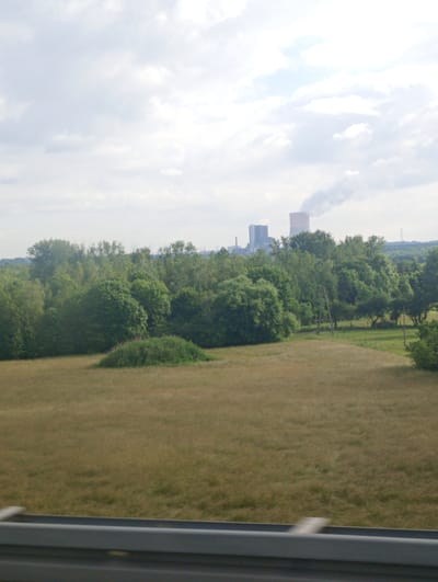 The image shows a landscape view with a power plant in the background. The power plant has two large structures, one of which is emitting a plume of smoke into the cloudy sky. In the foreground, there is a grassy field with some trees and bushes. A metal barrier is visible at the bottom of the image, suggesting the photo was taken from a vehicle or elevated position. The location is identified as Mysłowice, Poland.