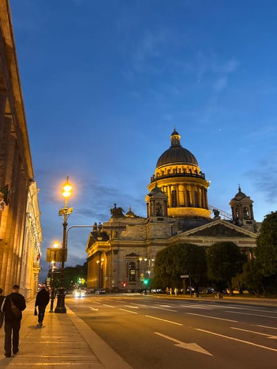 In the city of Unknown, Unknown, a majestic domed building, illuminated by warm light, dominates the scene. The sky above is a deep twilight blue, with wisps of clouds and a faint crescent moon visible. In the foreground, a street stretches out, lined with a sidewalk where several individuals walk. A streetlamp with two bright lights illuminates the sidewalk and a portion of the road. The building's facade features classical columns and ornate architectural details, with statues adorning its upper levels. Green trees are interspersed around the structure, adding a touch of nature to the urban landscape. Cars are visible on the road, suggesting the ongoing activity of the city. The overall atmosphere is one of serene evening grandeur.