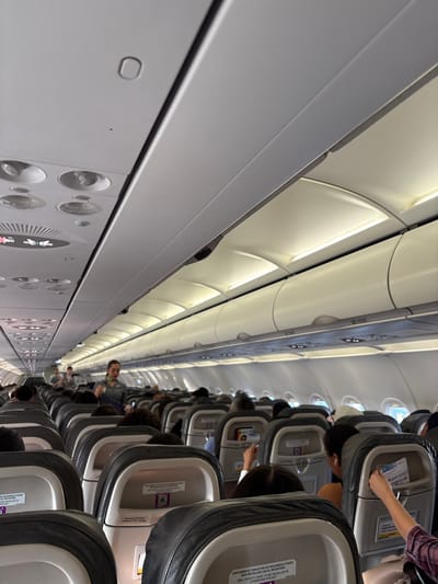 The image displays the interior of a commercial airplane cabin, viewed from an elevated and significantly tilted perspective, looking towards the rear of the aircraft. The main aisle runs diagonally across the frame, flanked by rows of passenger seats on both sides.

Numerous passengers are seated in the cabin, predominantly seen from behind, suggesting they are either awaiting departure or in transit. One individual in the foreground, seated on the right side of the aisle, is actively reaching into a seatback pocket. Other passengers further back are mostly obscured, though a few partial faces are visible.

The cabin features standard airline seating with dark grey and light grey elements. Overhead bins line the ceiling, which is illuminated by embedded lighting. The overhead panel on the left side of the image displays various controls and individual reading lights. Bright natural light filters in through the windows along the side of the plane, indicating daytime.

Visible text on a sticker attached to the back of a seat clearly states: "UNICAMENTE TARJETON DE SEGURIDAD PUEDE SER INSTALADO EN EL REVISTERO" and "ONLY SAFETY CARD TO BE INSTALLED IN LITERATURE POCKET." Additional partial text includes "DE SEGURIDAD," "EL REVISTERO," "BE INSTALLED IN POCKET," and "DAD PUEDE." The overall scene suggests a routine travel environment. The location context provided is Santa Cruz del Valle, Mexico.