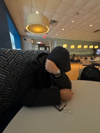 A person wearing a black jacket and hood sits at a table. The person's face is obscured by their arms. A watch is visible on their wrist. A phone connected to headphones sits on the table. In the background is a brightly lit cafeteria with tables and chairs. A wall with a sign reading "the BUSINESS of IMPROVING the WORLD" is visible. The person sits on a patterned couch. The image is rotated sideways.