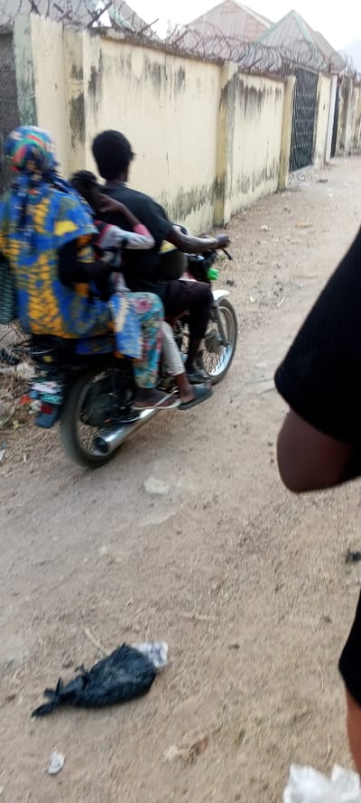 An unpaved dirt road in Nassarawa, Nigeria, serves as the setting for a daytime street scene. A motorcycle, moving from right to left, transports three individuals. The driver, an adult in dark clothing, operates the vehicle. A child in a light garment is positioned in front of a rear adult passenger. The rear passenger wears a vibrant blue, yellow, and multi-patterned traditional garment and a matching head covering, with feet in dark and white flip-flops. The motorcycle has a chrome exhaust and spoke wheels, with a bundle secured to the rear. To the right of the road, a weathered concrete wall topped with coiled razor wire extends along the street, featuring a dark metal gate further down. Building rooftops are visible beyond the wall. In the immediate foreground on the dirt road, a dark bag containing white material lies amongst scattered debris. The blurred arm and shoulder of an observer are partially visible in the lower right corner.