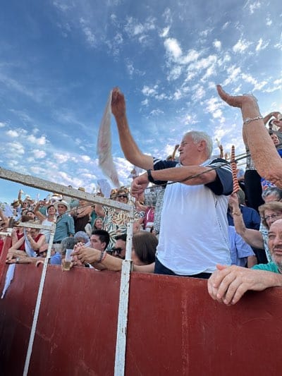 The image captures a vibrant scene in San José del Valle, Spain. Spectators are crowded behind the barrier of what appears to be a bullring, with the sky visible in the background. In the foreground, a middle-aged man with white hair is prominently featured, holding a white cloth aloft with his left hand. His right hand is also raised. The crowd appears to be cheering. The setting suggests an event or celebration related to Spanish culture.
