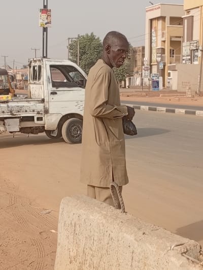 An elderly man in a beige tunic and trousers stands near a concrete barrier on a sandy road. He holds a dark bag in his left hand. A white utility truck is parked nearby. In the background are buildings, a street lamp with a poster, and glimpses of other vehicles on the road. The overall scene has a dusty, urban feel, possibly in a developing area.