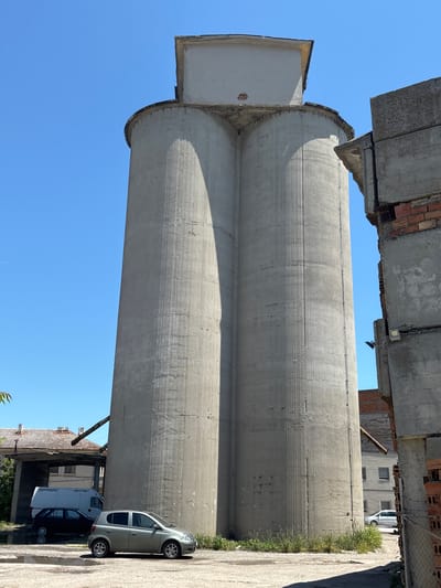 In Kavarna, Bulgaria, a towering concrete structure, likely a silo, dominates the frame under a clear blue sky. The silo appears to be comprised of two connected cylindrical sections, with a rectangular cap at its apex. Its weathered concrete surface bears vertical lines and subtle cracks. To the right, a partially constructed or dilapidated concrete and brick building stands, its exposed rebar and brickwork suggesting ongoing work or decay. In the foreground, a silver hatchback car is parked on a paved surface, with a black hatchback and a white van visible behind it. Vegetation sprouts around the base of the silo and the adjacent buildings, indicating an outdoor, potentially industrial or semi-abandoned, setting. The scene is illuminated by bright sunlight, casting distinct shadows.