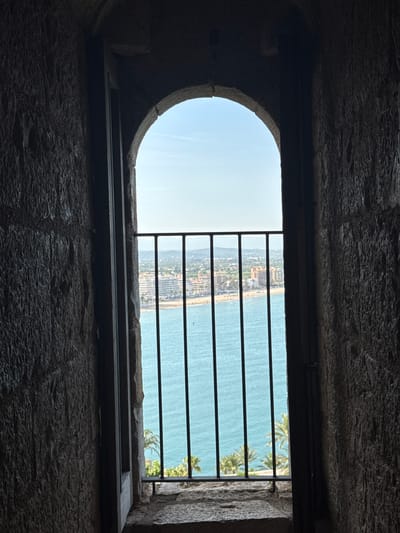 The image captures a scenic view from inside a dark, ancient stone structure, looking out onto a vibrant coastal landscape through a window opening. The window itself is framed by rough, dark stone walls and a thick sill, with a distinctive rounded arch on its left side. Several horizontal black metal bars are positioned across the opening, suggesting a protective or fortress-like setting.
Beyond the window, a bright, clear blue sky dominates the upper left. The main view showcases a wide expanse of turquoise-blue sea on the right, meeting a long, sandy beach. Along this coastline, numerous buildings form a dense urban strip, indicating a populated town or resort area. Details of these structures and some green vegetation are visible along the coast. On the far right, behind the metal bars, the fronds of palm trees are discernible, suggesting a warm, Mediterranean climate.
The scene is bathed in bright daylight, implying a clear, sunny day, likely mid-day or early afternoon. The strong contrast between the shadowy interior and the brightly lit exterior draws the viewer's eye towards the distant landscape. The setting, with its ancient stone architecture and coastal panorama featuring a populated beach, strongly aligns with the provided context of Peníscola, Spain, renowned for its historic castle overlooking the Mediterranean Sea. There is no visible text in the image.
