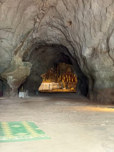 This is a medium shot, eye-level, indoor photograph of the interior of a cave. The cave has rough, textured, gray-brown rock walls and a sandy floor. In the foreground, a green and yellow woven mat is placed on the floor. The cave opens into a larger chamber in the background, where numerous small, golden Buddha statues are displayed on shelves. Some of these statues appear to be illuminated by artificial lighting, casting a warm glow. The cave is a place of worship. It is located in Xanghay, Laos.